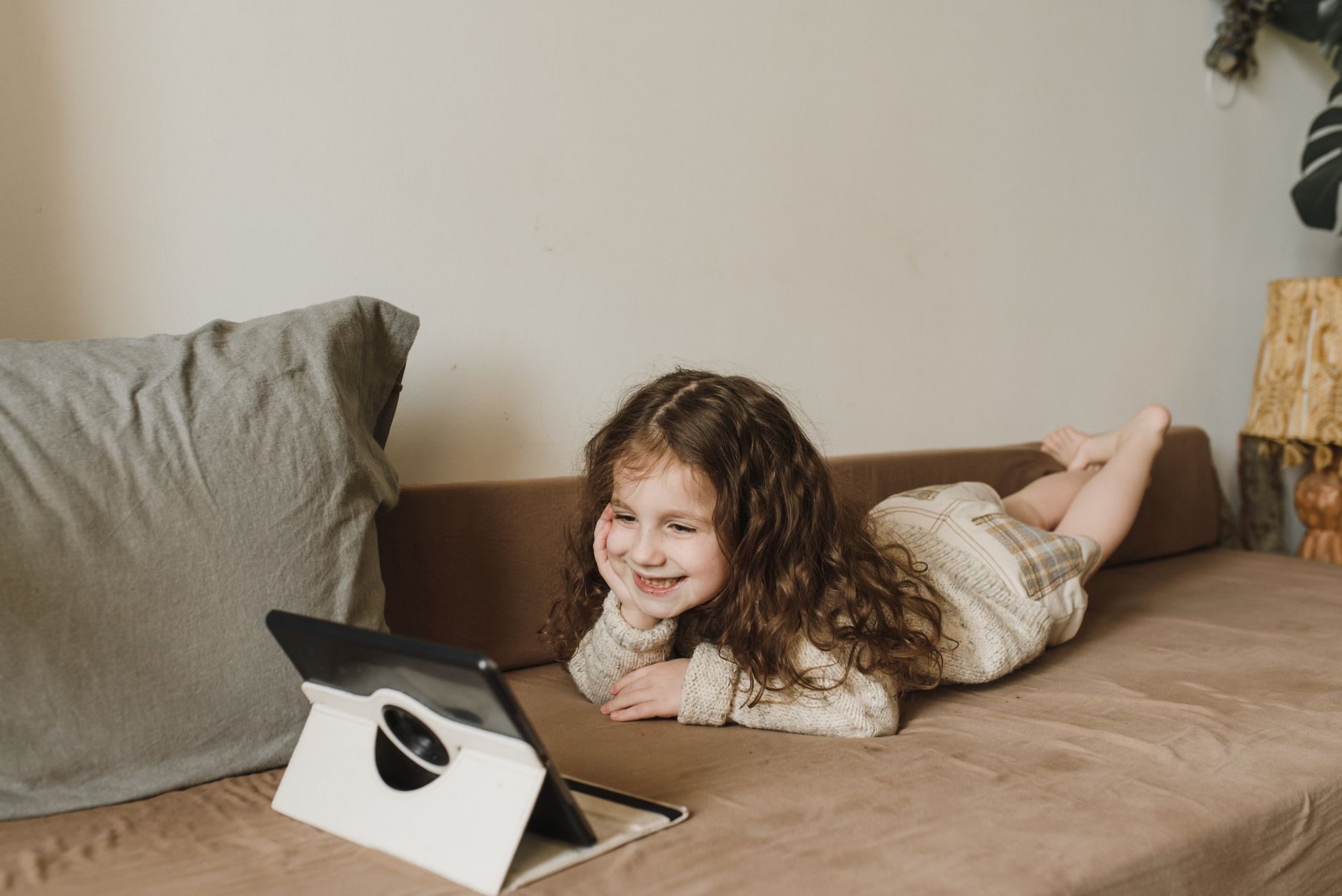 Jeune fille allongée sur un canapé, regardant joyeusement une tablette dans un intérieur confortable.