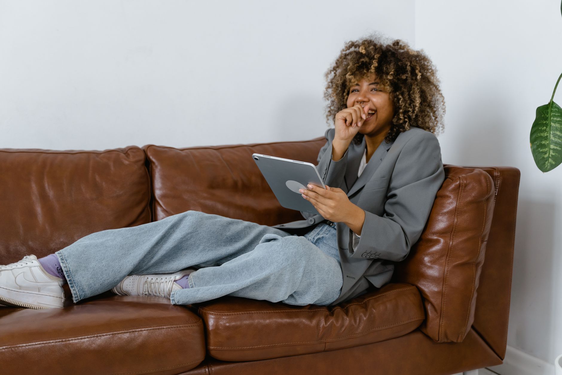 Femme riant en utilisant une tablette, installée sur un canapé en cuir marron.
