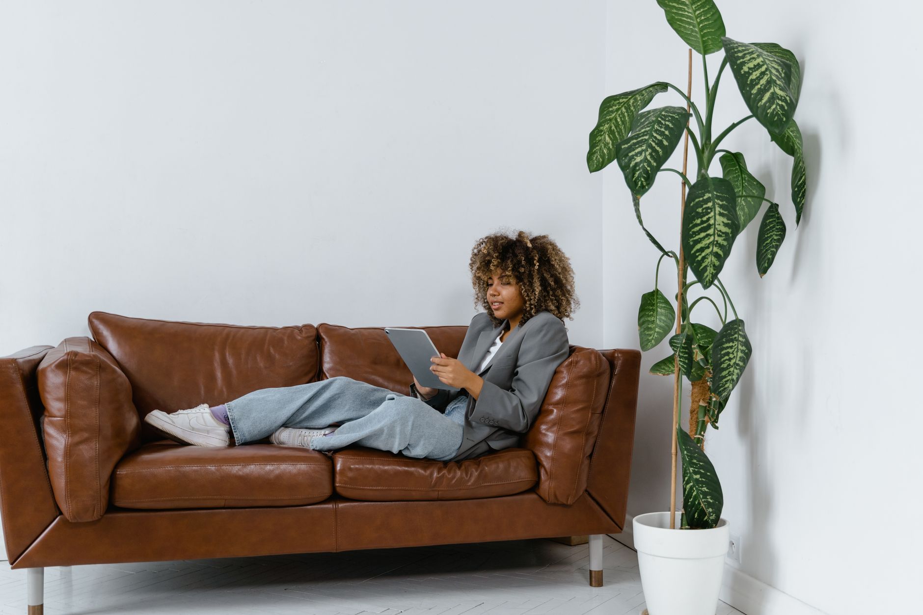 Femme installée sur un canapé en cuir, tablette en main, près d'une plante verte.